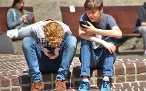 Two boys sit outside together on their phones