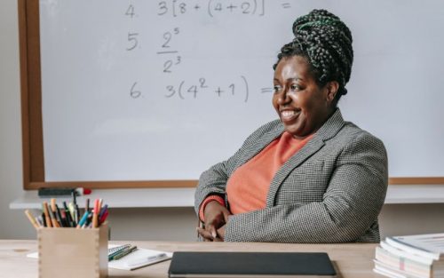 A female teachers sits at her desk smiling with a whiteboard full of maths problems behind her