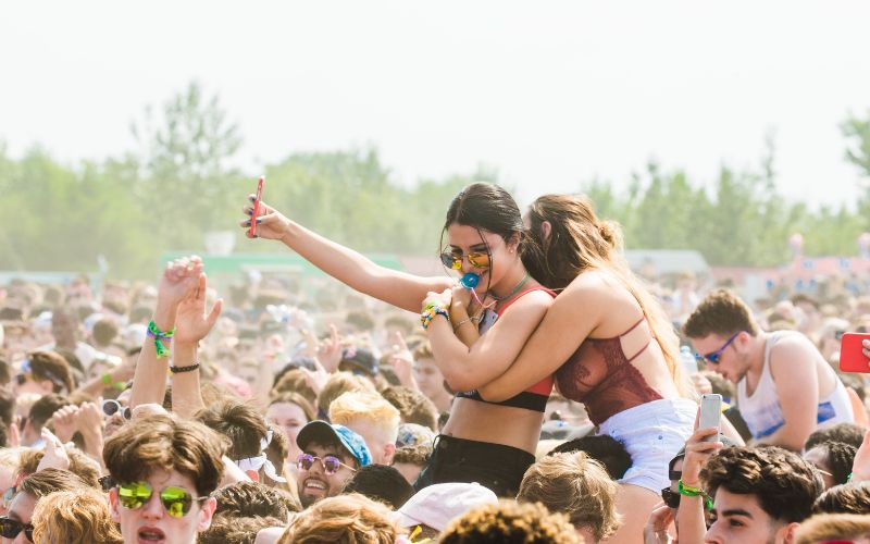 Two young women sit on the shoulders of their friends at a music festival, they are filming music acts with their phones